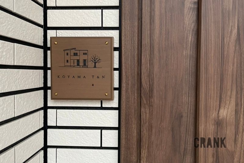 白いタイル壁の角に設置された、正方形の木製表札「KOYAMA T&N」。表札にはご自宅の外観と桜の木のイラストが彫刻されており、右隣には落ち着いた木目の玄関ドアが見えている。