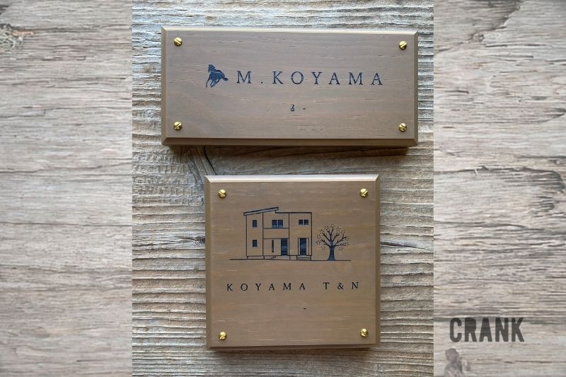 木目の背景の上に並べられた2種類のウォルナットカラーの木製表札。上には馬のモチーフがあしらわれた横長の表札、下にはご自宅の外観と桜の木が描かれた正方形の表札。それぞれ四隅がゴールドの真鍮ネジで留められている