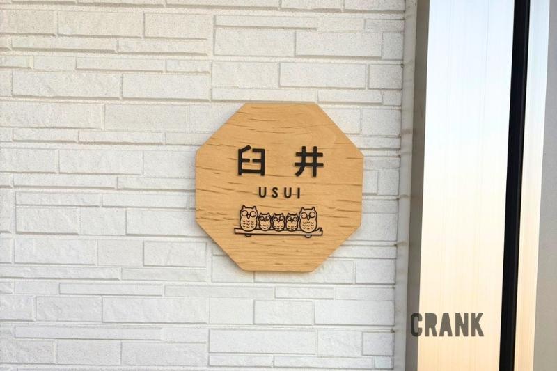白いタイル調の外壁に設置された、八角形の木製表札「臼井」。文字の下には、木の枝に仲良く並んだ5匹の親子フクロウのモチーフが彫刻されている。下部にはシルバーのインターホン。